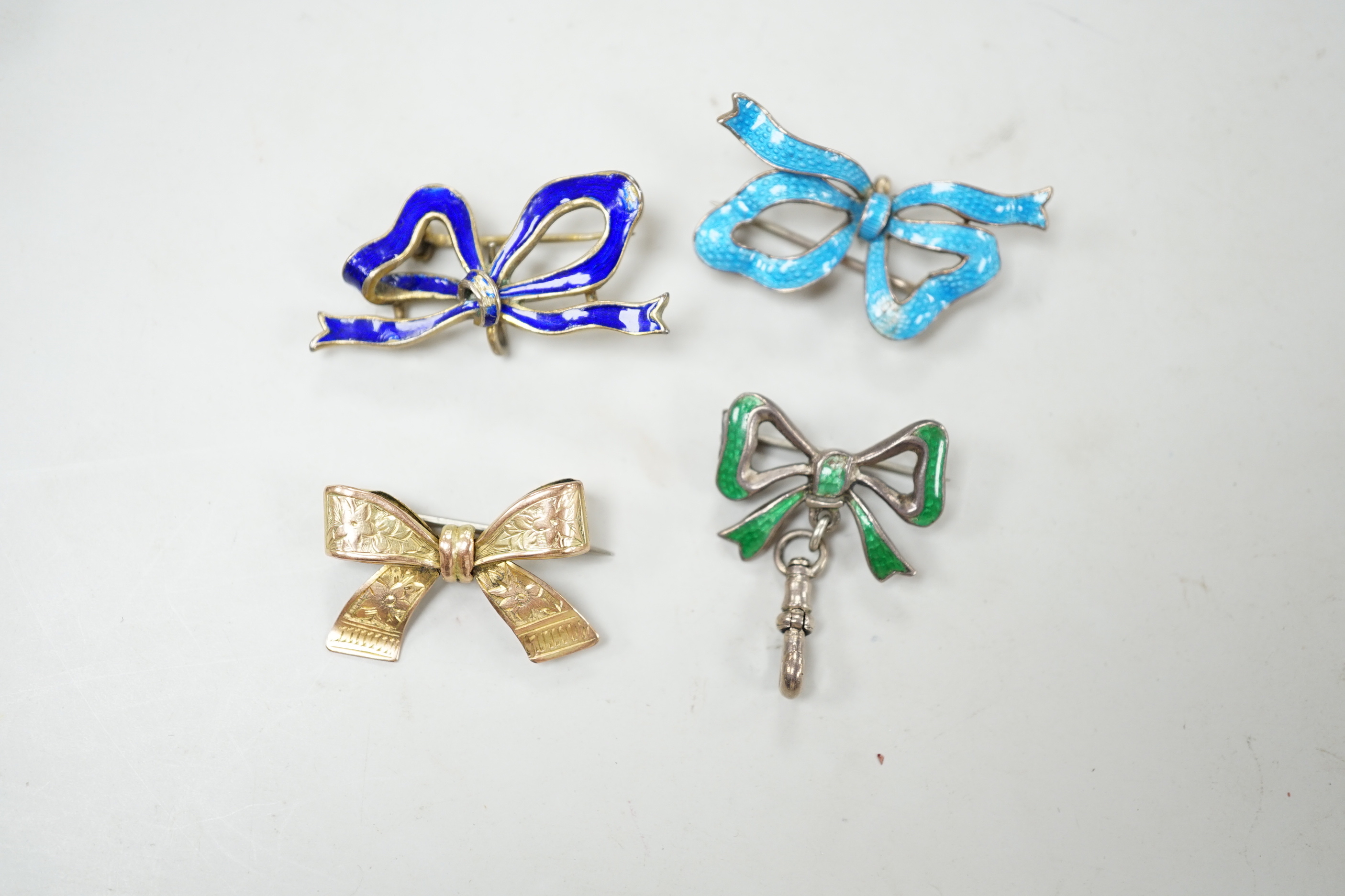 An engraved yellow metal ribbon bow brooch, 26mm, 2.8 grams, together with three silver/gilt white metal and enamel ribbon bow brooches.